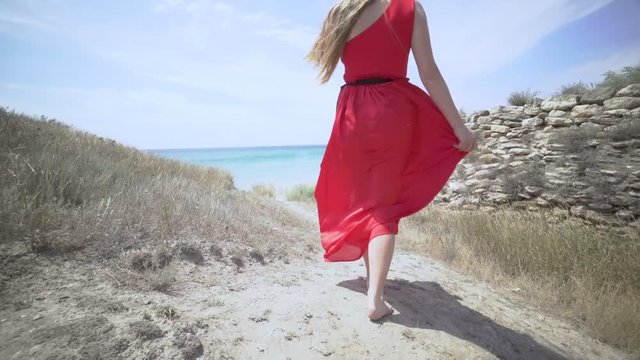 Young girl in red dress walking barefoot on a path towards the sea. Girl's long hair and bright scarlet gown blowing in the wind. Windy summer day at the seaside. Gimbal steadicam shot.