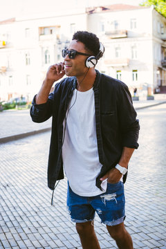 Portrait of young stylish hipster black man in white headphones and sun glasses dancing outdoor in city centre, having some fun