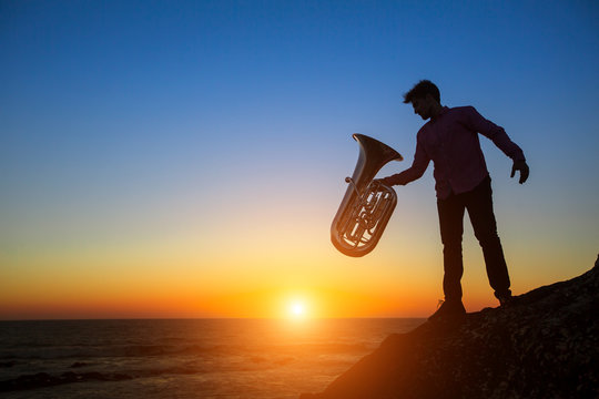 Silhouette of musician with the Tuba on rocky sea coast during sunset.