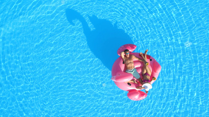AERIAL TOP DOWN: Young couple drinking cocktails and relaxing while lying on fun inflatable pink flamingo floatie. Sexy couple in swimsuit sipping drinks and enjoying floating on sunny pool water © helivideo