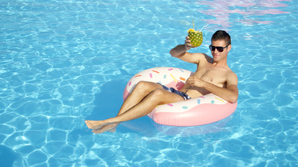 CLOSE UP Happy young guy on summer vacation lying on inflatable doughnut floatie in swimming pool & drinking pineapple alcohol beverage. Smiling man floating on inflatable pillow and sipping cocktail - Powered by Adobe