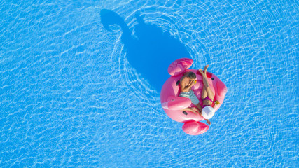 AERIAL TOP DOWN: Young man and woman drinking cocktails and relaxing while lying on fun inflatable pink flamingo floatie. Sexy couple in swimsuit sipping drinks, enjoying floating on sunny pool water