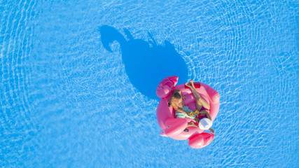 AERIAL TOP DOWN Young couple drinking cocktails and relaxing while lying on fun inflatable pink flamingo floatie. Sexy boyfriend and girlfriend sipping drinks and enjoying floating on sunny pool water