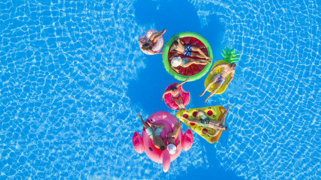 AERIAL TOP DOWN COPY SPACE: Fit girls and muscular guys lying on colorful floaties in pool, playing volleyball with a ball. Friends on fun summer vacation floating on water on inflatable loungers"