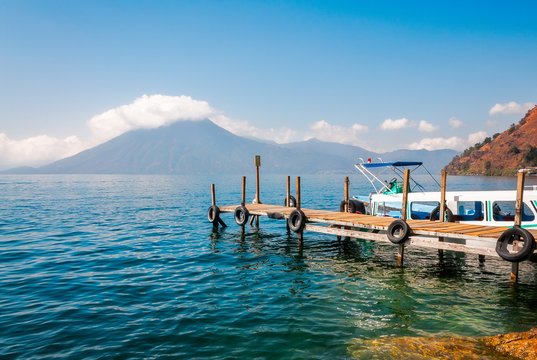 Astounding View of Volcano San Pedro with a crown of clouds from the shore of Lake Atitlan in Guatemala with a tourist boat docked at a pier.