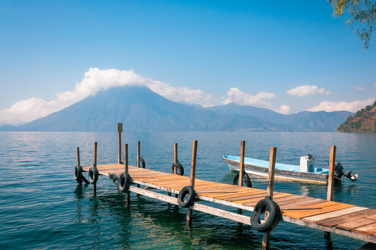 Astounding View of Volcano San Pedro with a crown of clouds from the shore of Lake Atitlan in Guatemala with a tourist boat docked at a pier.