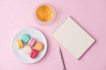 Flatlay of notebook, cake macaron and cup of tea on pink table. Beautiful breakfast with macaroon.