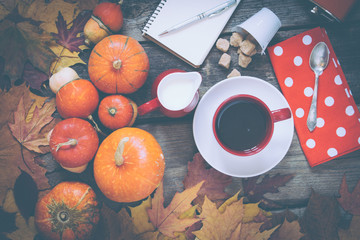 red cup of tea or coffee, milk jug, notepad and pen, red napkin at polka dots on wooden table decorated by falling leaves and pumpkins, top view