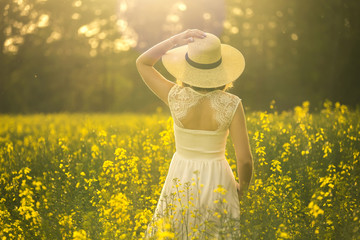 Portrait of young and attractive woman in white dress and with hat in rape field. Soft focus. Backside view.