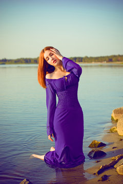 Gorgeous sexy young lady at evening sunlights around rocks and water. Woman wearing in purple long dress. Portrait in vintage style. Sweet woman rest on the sea