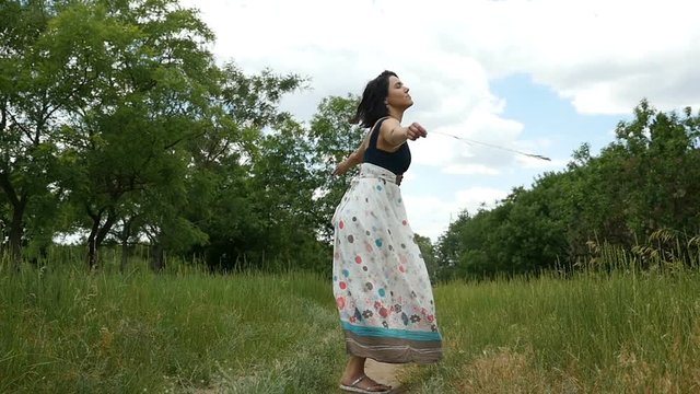 A blissful brunette woman in a folk dress with open shoulders turns around on a country road and smiles happily in slow motion. She is on a green  lawn with green trees on a sunny day in summer