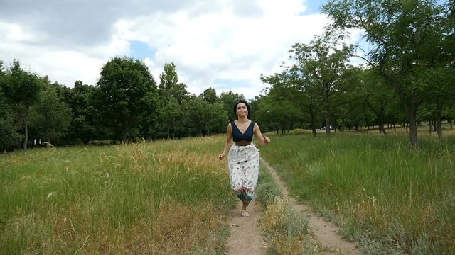 A charming brunette woman, in a white dress with black top and open shoulders runs along a country road and smiles happily in slow motion. She is on picturesque lawn with green trees on a sunny day