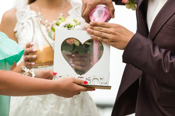 Groom and bride pouring colourful sand into box with glass heart outdoor