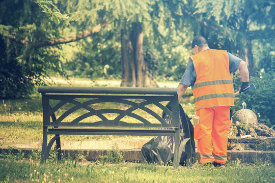 sweeper working at the park, person with high visibility vest collects garbage near a bench