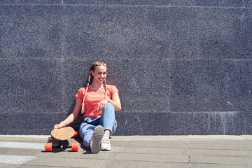 Gorgeous female while sitting under wall with skateboard
