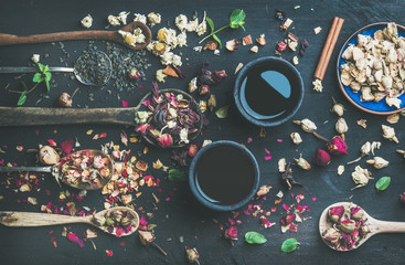 Chinese black tea in black stoneware cups and wooden spoons with dry herbs, flower buds and leaves over black wooden background, top view, horizontal composition
