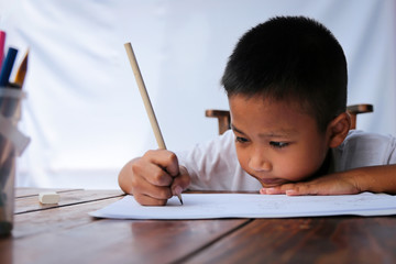 Asian little boy drawing intently on wooden table with sunlight and white background. Little painter concept.