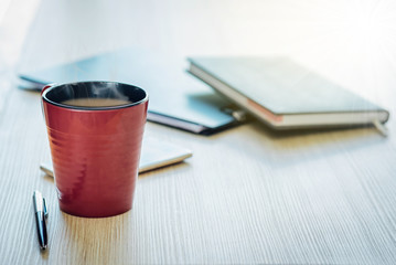 The reddish brown coffee cup is on the wood table and pen, the diary book is blurred background.