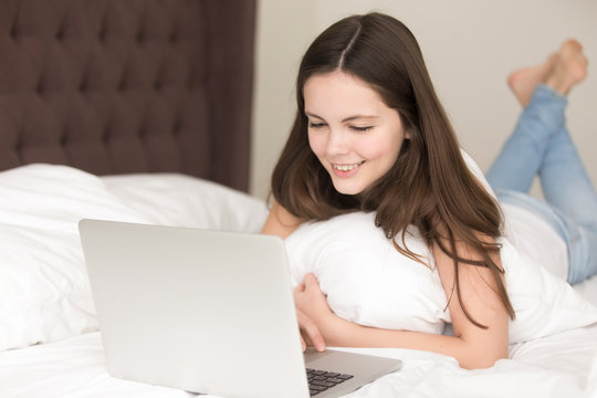 Pretty young lady using laptop while lying on bed in the morning, smiling teen girl checking news feed on pc at home, playing games, browsing web, shopping online, communicating with friends