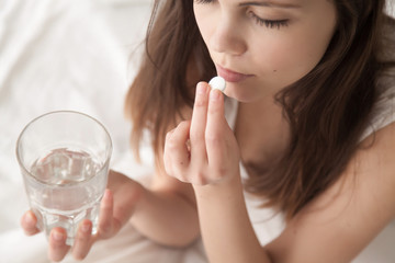 Unhealthy sick woman suffers from insomnia or headache, takes sleeping pill while sitting in bed with glass of water, depressed girl holds antidepressant meds, painkiller for menstrual pain, close up