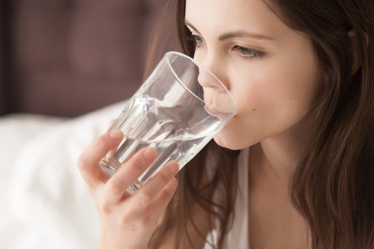 Attractive young dehydrated woman feeling thirsty, holding glass and drinking pure mineral water every morning after waking up, good habits for healthy way of life, dehydration concept, close up view