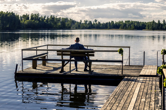 Portrait of young and attractive man in suit sitting on a bench on pier next to the huge lake and enjoying the view.