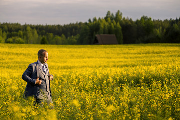 Young and attractive man in a suit standing in yellow rape field and looking somewhere.
