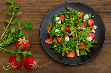 Salad with arugula, strawberries, cottage cheese, olive oil, on a black plate, wooden background. Close-up. Top view