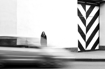 Girl standing against a white wall with a black and white gates. Ride car in motion blur in the foreground.