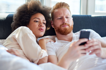 Young couple laughing together and using mobile phone while lying in bed