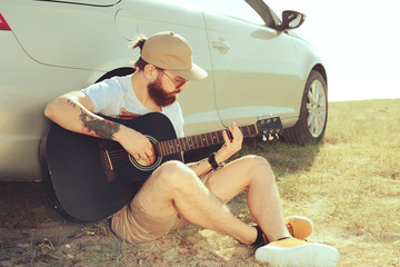 Bearded man playing guitar outdoors near car