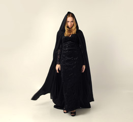 Naklejka premium full length portrait of a blonde lady wearing black lace now and hooded cloak, standing pose isolated against creamy background.