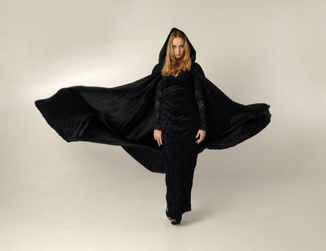full length portrait of a blonde lady wearing black lace now and hooded cloak, standing pose isolated against creamy background.