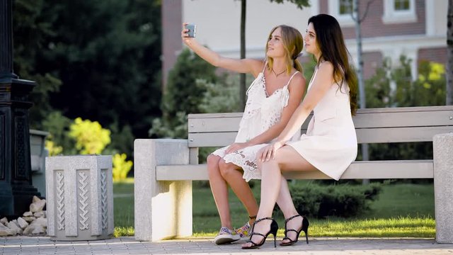 Lovely girls are sitting on a bench in by near the house, girl takes selfies and her friend happily chatting