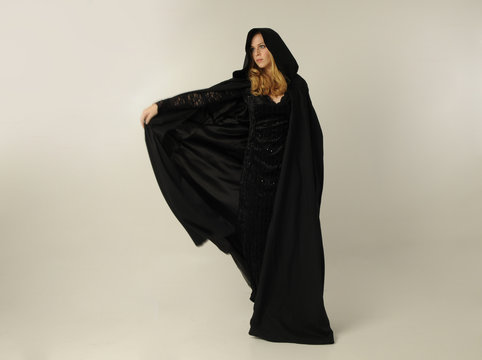 full length portrait of a blonde lady wearing black lace now and hooded cloak, standing pose isolated against creamy background.