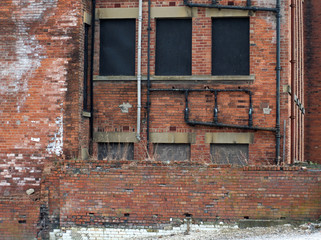 derelict abandoned commercial or factory type building with boarded up windows decaying bricks and a collapsing wall with weeds