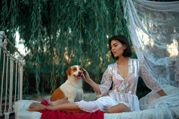 Young sexy woman with dog sits on bed with bedding and baldachin near tree. © Stanislav