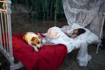 Young woman with dog lies on bed with bedding and baldachin near tree and sleeps. © Stanislav