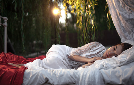 Young pensive woman lies on bed with bedding and baldachin near tree on sunset.
