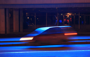 The car, lit in blue, travels through the evening city with a blur in motion 