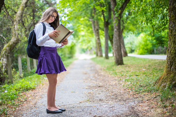Pretty beautiful blonde child girl reading book standing back to school enjoying through alley in the nature with backpack, glasses, purple skirt and white shirt