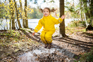 Cute little girl jumping in muddy puddle wearing yellow rubber overalls. Happy childhood. Sunny...