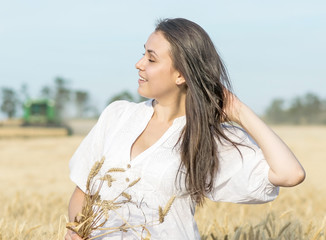 Beautiful sexy female touching her long dark hair in wheat field during harvesting. Harvester in the back blurry background
