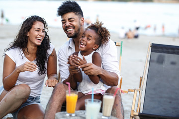 Young mixed race family sitting and relaxing at the beach on beautiful summer day.Daughter lies in father lap and laughing.