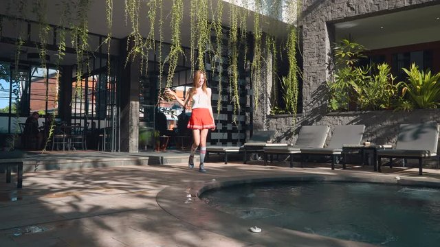 Young caucasian woman in sport costume (white top, red skirt, football stockings and sneakers) walks to swimming pool edge and stretches out arms in the morning . Shot with Sony a7s.