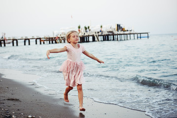 Cute little girl in pink dress running and dancing on sand at seashore. Vacation and holidays concept. 