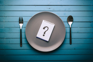 Empty plate with a question mark drawn on a notepad on a blue table, food concept