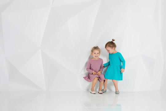 Two little girlfriends in the identical dresses of different colors sitting in a studio with white walls