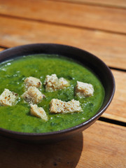 Green spinach soup with piece of bread in brown bowl on wooden table.