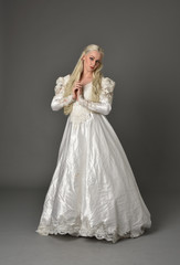 full length portrait of a beautiful blonde woman wearing link white gown, standing pose against a grey background.